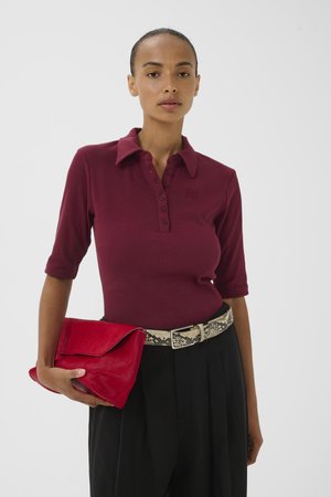 Frau trägt ein burgunderfarbenes Poloshirt mit Knöpfen, schwarze Hose mit einem Schlangenledergürtel und hält eine rote Clutch vor einfarbigem Hintergrund.