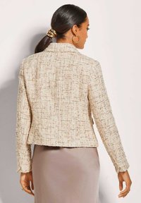 Beige tweed jasje met gestructureerde weving, getailleerde snit en kenmerken van een notch-kraag. Lange mouwen met knoopmanchetten. Draagt over een glad, licht rok.