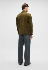 Olive green knit sweater with textured stripes, high collar, and ribbed cuffs, paired with loose blue denim jeans, viewed from the back.