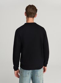 Maglione nero lavorato a maglia con maniche lunghe, scollo tondo e polsini e orlo a coste. Indossato con jeans azzurri, che mostrano una vestibilità comoda.