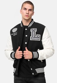 Chaqueta de varsity en blanco y negro con parches bordados, botones frontales, puños acanalados y dobladillo a rayas; tela suave con mangas en contraste.