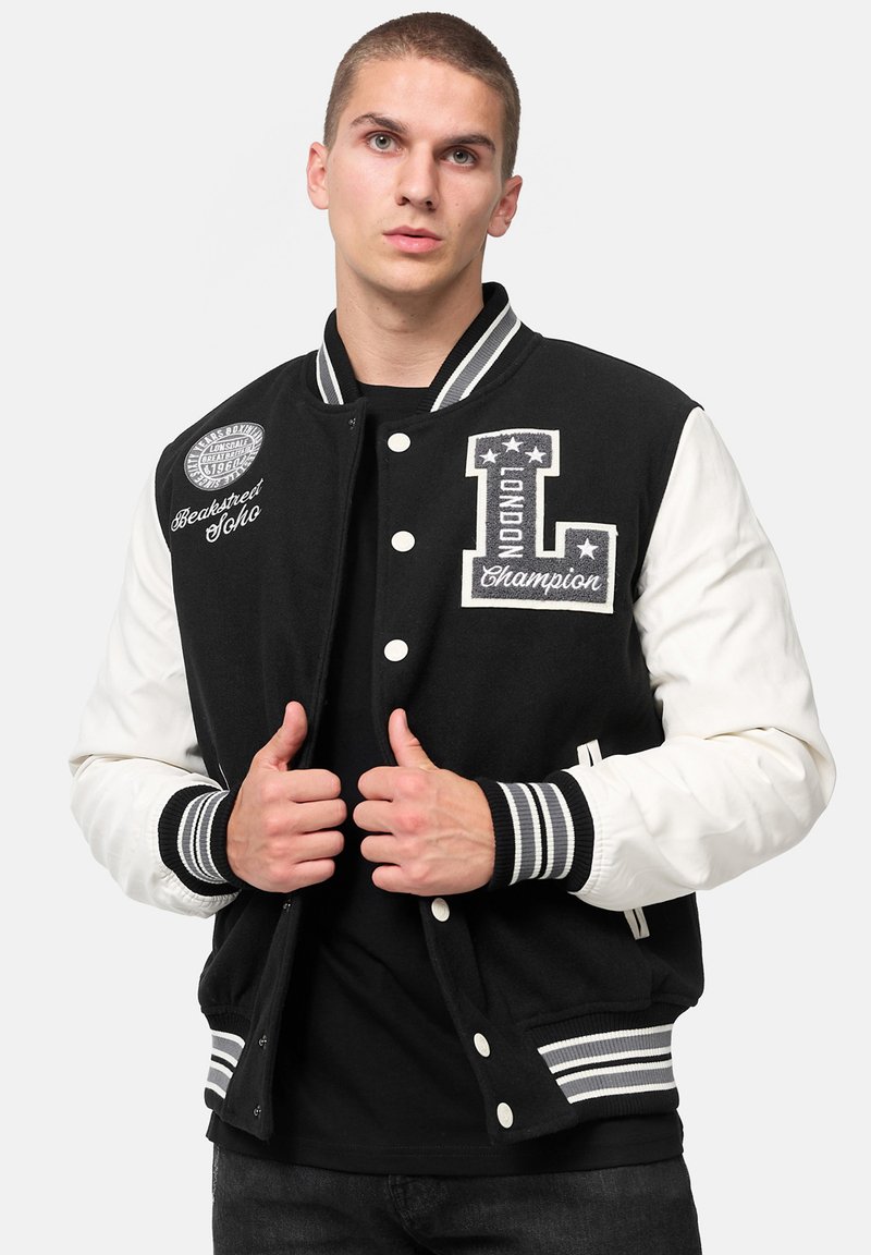 Chaqueta de varsity en blanco y negro con parches bordados, botones frontales, puños acanalados y dobladillo a rayas; tela suave con mangas en contraste.