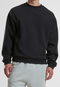 Homme portant un sweat-shirt noir uni à manches longues et col rond, accompagné d’un pantalon gris clair, debout devant un fond neutre.