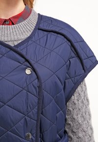 Navyblå quiltad väst med bred axeldesign, silverfärgade tryckknappar och ribbad grå krage över en mönstrad grå tröja.