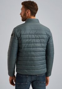 Giacca imbottita di blu tenue, con linee trapuntate orizzontali, colletto alto e tasche laterali. Indossata su jeans di denim.