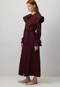 Burgundy lång klänning med volanger vid krage och manschetter, knappfront och midjeband. Slät tyg med svävande silhuett. Bärs med beige skor.
