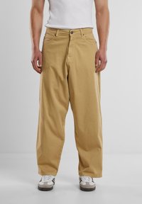 Beige cotton wide-leg pants with a button closure, two front pockets, and a relaxed fit, paired with white sneakers for a casual look.