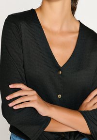 Blouse noire texturée avec un décolleté en V et trois boutons. Présente des manches fluides et une coupe générale décontractée.