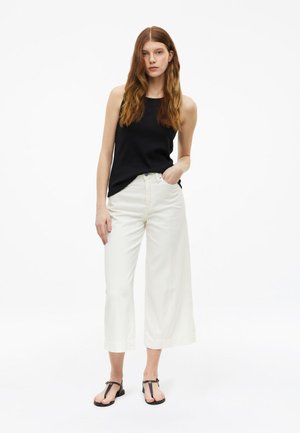 Junge Frau mit langen roten Haaren, die ein schwarzes ärmelloses Top, weiße weite Cropped-Hosen und schwarze Sandalen trägt, steht mit einer Hand in der Tasche.