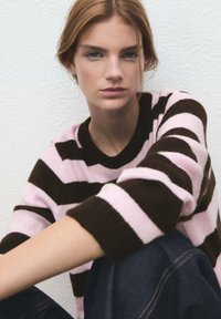 Jersey de punto a rayas con franjas rosas y marrones. Presenta un escote de cuello redondo y mangas enrolladas. Usado sobre jeans oscuros. Tejido texturizado.