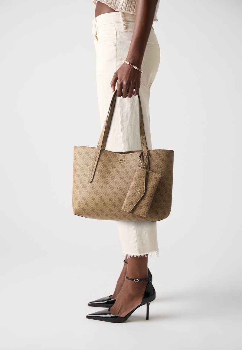 Sac fourre-tout en monogramme beige fabriqué dans un matériau texturé, doté de deux longues poignées et d'un portefeuille détachable. Porté avec un pantalon crème et des talons noirs.