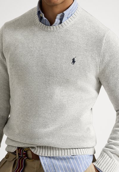 Svetlo siva pletena majica z nazobčanim okroglim ovratnikom in majhnim temnomodrim logotipom na prsih. Nosi se čez modro srajco s črtami.