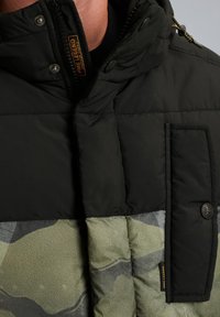 Detailný záber na čiernu a zelenú maskáčovú prešívanú bundu s vreckom na klopu so zapínaním na gombík a štítkom PME Legend na hrudi.