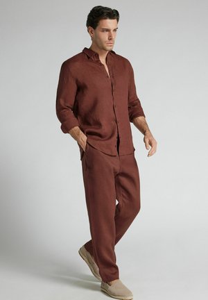 Man met donker haar, gekleed in een bruine lange mouwen blouse met knopen en bijpassende broek, lopend met handen in de zakken, op een lichtgrijze achtergrond.