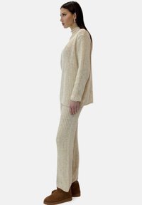 Roomkleurige geribbelde trui en bijpassende broek in zachte stof, met een hoge halslijn en een ontspannen pasvorm, gecombineerd met bruine enkellaarzen.