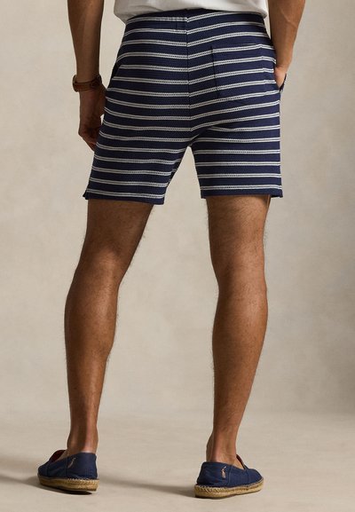 Mann som står med hendene i lommer, iført marineblå og hvite stripete strikkeshorts og marineblå espadrillosko mot en ensfarget bakgrunn.