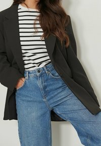 Blazer noir porté sur une chemise rayée blanche et noire, associé à un jean taille haute bleu clair. Le tissu semble lisse avec un design structuré.