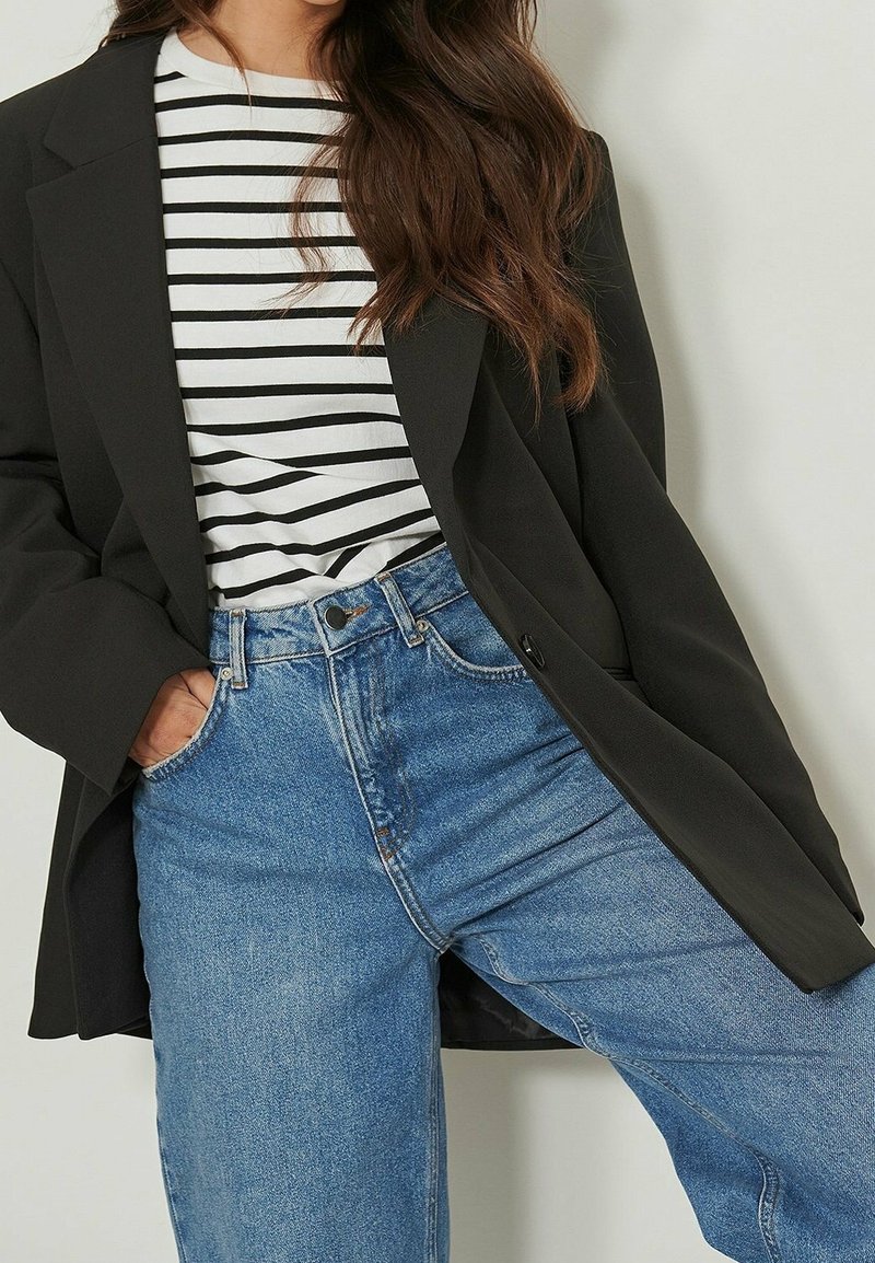 Blazer noir porté sur une chemise rayée blanche et noire, associé à un jean taille haute bleu clair. Le tissu semble lisse avec un design structuré.