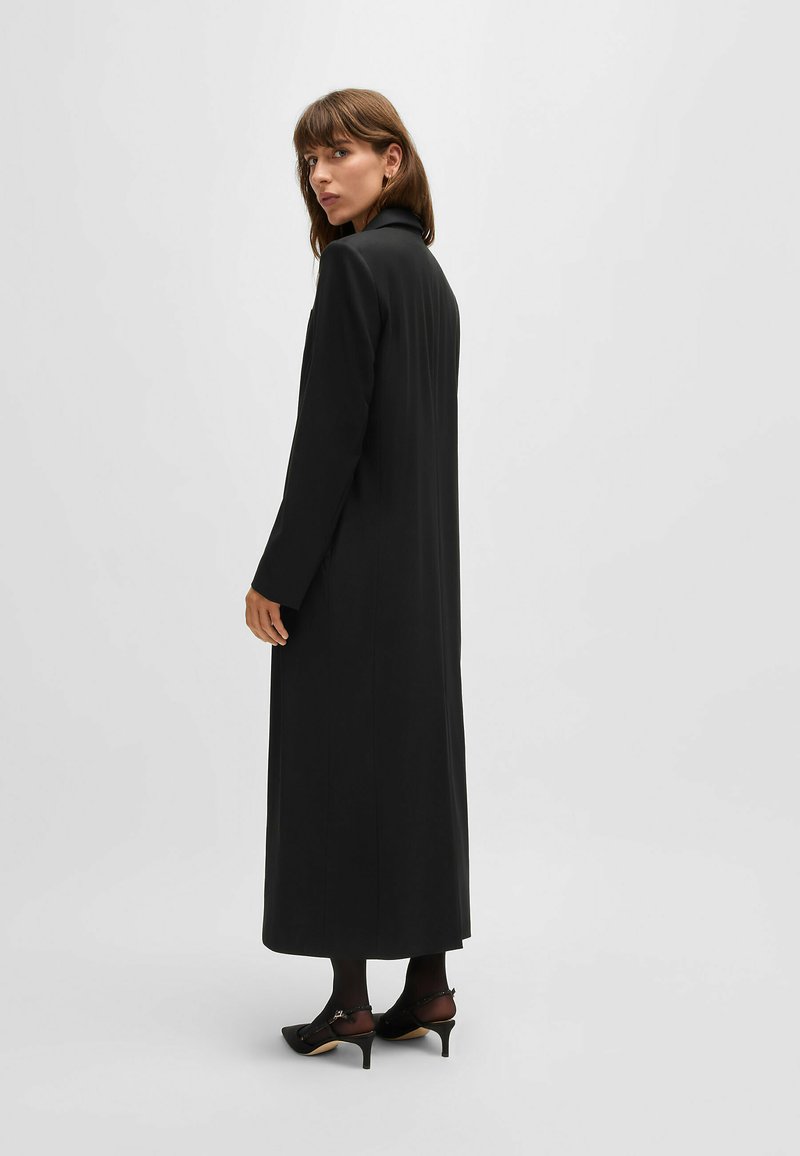 Manteau long noir avec un col montant, coupe droite et manches longues, associé à des bas noirs et des talons aiguille avec des brides de cheville.
