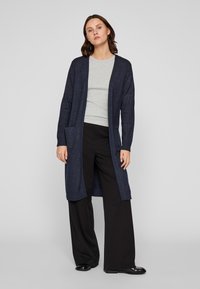 Μακριά μπλε κάρντιγκαν φτιαγμένο από υφασμάτινη υφή, με ίσια γραμμή, ανοιχτό μπροστά, δύο μπροστινές τσέπες και πλεκτές μανσέτες. Φορεμένο πάνω από ένα γκρι τοπ.