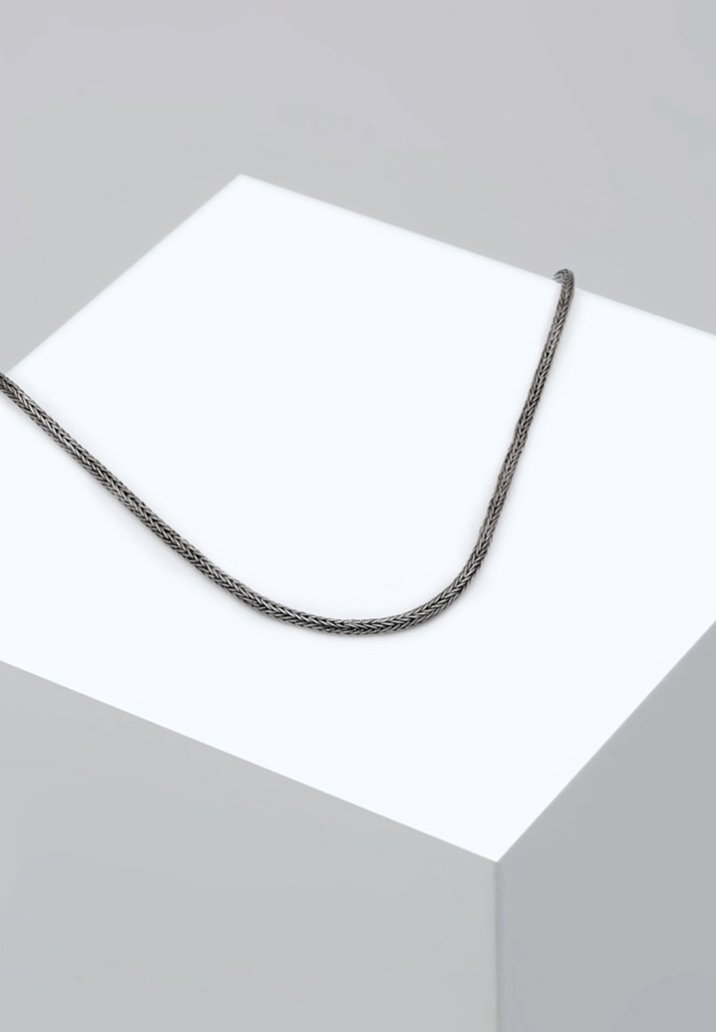 Silverflätat kedjehalsband som vilar på en vit piedestal, visar en slank, rundad form och en slät yta med en minimalistisk design.