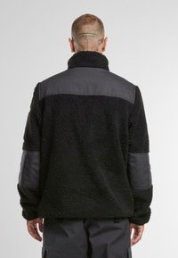 Giacca di fleece nera in materiale testurizzato e morbido. Presenta un pannello grigio sulle spalle, con accenti laterali eleganti e un colletto alto.