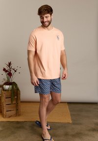 Perzikkleurig katoenen T-shirt met logo, gecombineerd met blauw patroon zwembroek. Lichte schoeisel, staand op een gestructureerde vloer met natuurlijke decoratie.