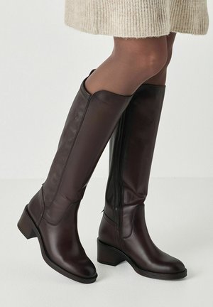 Botas de cuero marrón oscuro hasta la rodilla, usadas con medias marrones translúcidas y una falda beige de punto sobre un fondo claro.