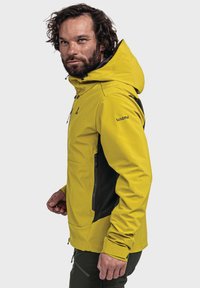 Gelbe wasserdichte Jacke mit einem schwarzen Seitenpanel, verstellbarer Kapuze, Frontreißverschluss und Seitentaschen. Glatte Textur und sportlicher Schnitt.