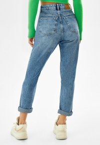 Vaaleansiniset korkeavyötaröiset farkut, joissa on kapeneva malli, käänteet lahkeissa ja kaksi takataskua, yhdistettynä kermaiset tennarit, joissa on kultaketju yksityiskohta.