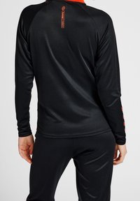 Camiseta deportiva de manga larga en negro, con un color y logotipo en rojo, fabricada con un tejido texturizado y un diseño aerodinámico que facilita el movimiento.