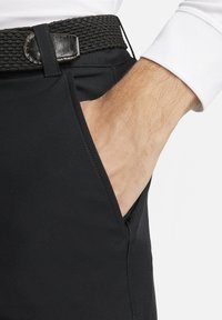 Juodos elastinės kelnės turi šoninę kišenę ir pintą juodą diržą su odiniu akcentu. Artimas vaizdas pabrėžia tekstūrą ir detales.