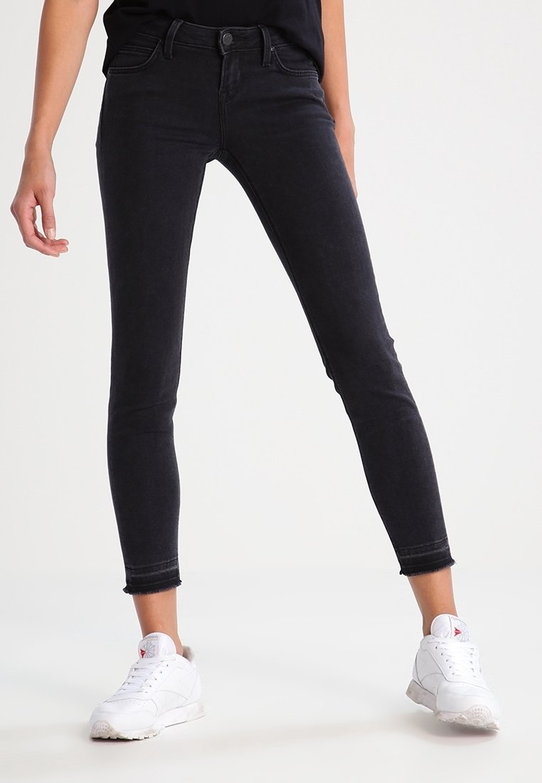 Jeans ajustados negros con una textura suave, cintura de altura media, detalles de desgarro sutil y ligero deshilachado en el dobladillo, combinados con zapatillas blancas.