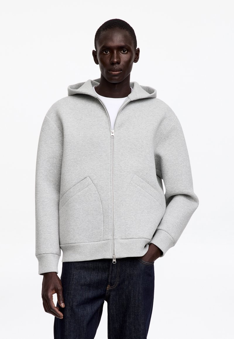 Homme portant un sweat à capuche zippé gris clair avec capuche et poches avant, associé à un jean bleu foncé, debout devant un fond uni.