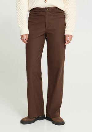 Persona che indossa pantaloni marroni a gamba dritta, cintura marrone intrecciata, maglione beige strutturato e scarpe marroni, in piedi su uno sfondo chiaro.