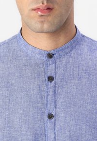 Kék chambray ing, kerek gallérral, három fekete gombbal és texturált anyaggal, a nyakkivágás körüli közeli nézetben.