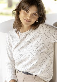 Jeune femme aux cheveux bruns mi-longs sourit, portant un haut blanc à pois, un pantalon beige et des boucles d'oreilles créoles dorées, appuyée sur une surface blanche.