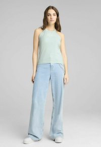 Lichtgroene haltertop gecombineerd met losse lichtblauwe jeans, met een kettingaccent op de zak, en witte sneakers. Effen achtergrond.