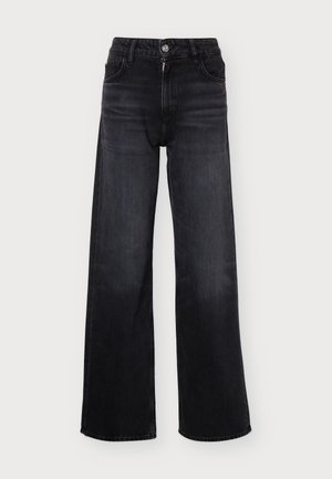 Un mannequin se tient devant un fond uni clair, vêtu d’un col roulé noir, d’un jean noir à jambes larges et de sandales ouvertes.