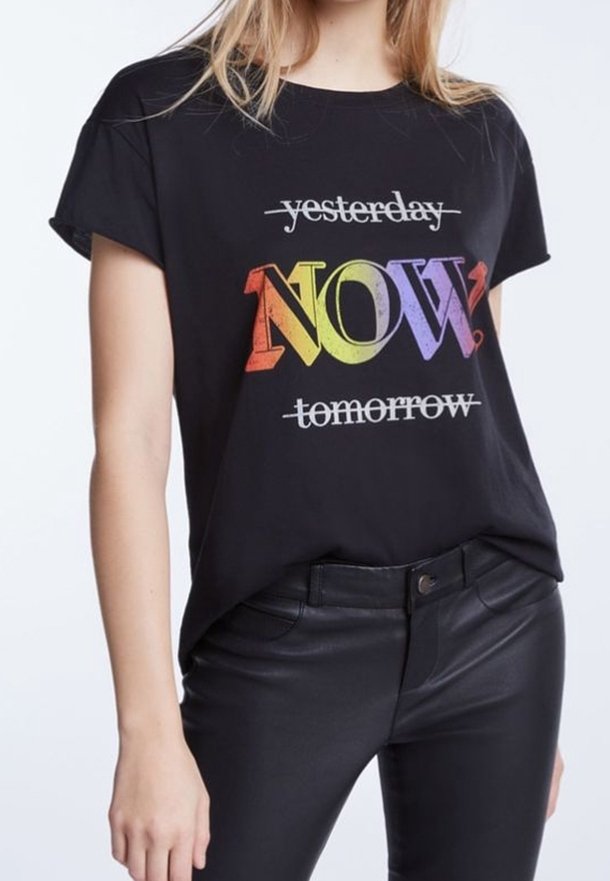 Femme portant un t-shirt noir avec les mots "hier" et "demain" barrés et le grand mot coloré "MAINTENANT ?" au centre.