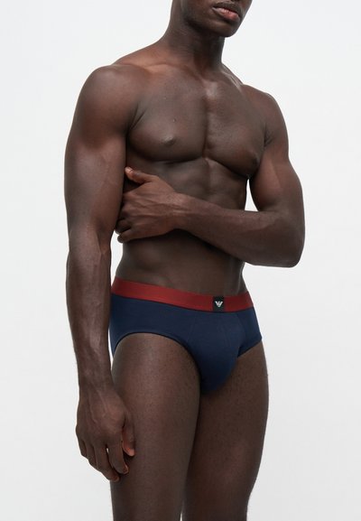 Homme musclé à la peau foncée portant un slip bleu marine avec une ceinture rouge, debout devant un fond clair uni, main sur le côté du torse.
