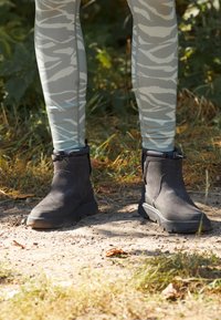Schwarze Wildleder-Ankle Boots mit Gummisohlen, ausgestattet mit einem verstellbaren Knöchelriemen, kombiniert mit grauen gemusterten Leggings in einer natürlichen Umgebung.