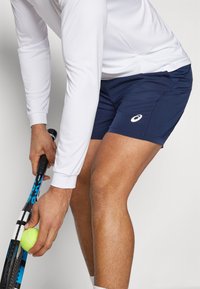 Marineblå sportshorts lavet af letvægtsstof, med en sidelomme og et lille hvidt logo. En hånd holder en tennisracket og en bold.