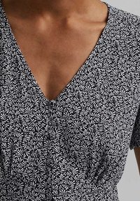 Schwarzes Kleid mit einem weißen Blumenmuster, V-Ausschnitt, kurzen Ärmeln und gerafftem Bund. Der Stoff wirkt leicht und hat eine glatte Textur.