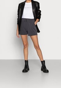 Grijze shorts met een elastische tailleband en koord, gecombineerd met een wit shirt en een zwarte blazer. Zwarte enkelboots maken de outfit compleet.