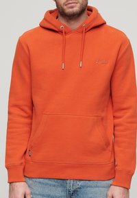 Sudadera con capucha de algodón naranja con bolsillo frontal, cordones ajustables y puños acanalados. Presenta un logo sutil en el pecho.