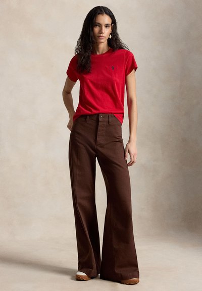 Frau steht mit einer Hand in der Hüfte und trägt ein rotes Kurzarmhemd, braune weite Hosen, weiße Schuhe und einen großen Ohrring vor einem einfarbigen Hintergrund.