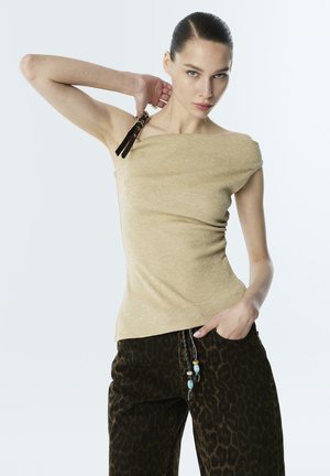Vrouw met naar achteren gestyled haar draagt een beige top met één schouder en een zwarte gespband, en donkere broek met luipaardprint en een koord met kralen, poserend met hand in de zak.