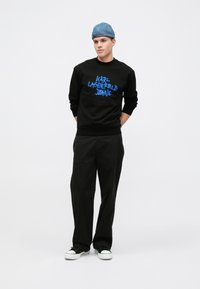Svart hettegenser med blå tekst "KARL LAGERFELD JEANS," kombinert med svarte vide bukser og svarte joggesko med hvite såler. Denim-kaps.