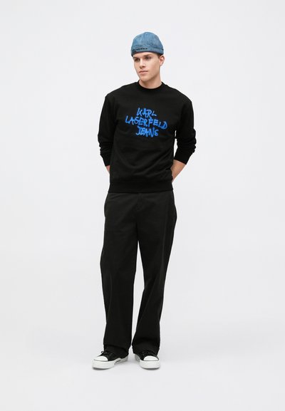 Sudadera negra con texto azul "KARL LAGERFELD JEANS", combinada con pantalones anchos negros y zapatillas negras con suelas blancas. Gorro de mezclilla.